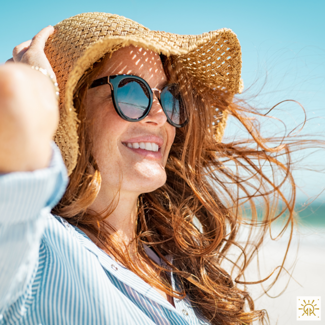 Red headed girl with sunglasses holding onto her hat on the beach